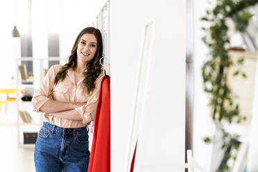 Smiling female fashion designer with arms crossed leaning on rack at studio - GIOF09810