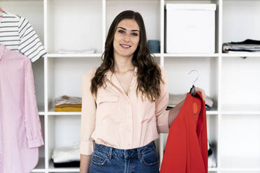 Smiling female designer holding red winter coat while standing against rack in studio - GIOF09812