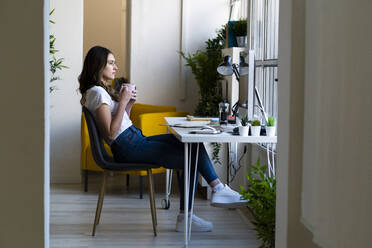 Thoughtful businesswoman holding coffee cup while sitting on chair at office - GIOF09813