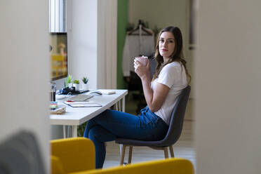 Geschäftsfrau hält Kaffeetasse, während sie auf einem Stuhl im Büro sitzt - GIOF09814