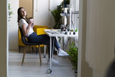 Lächelnde Geschäftsfrau, die eine Kaffeetasse hält und auf einem Stuhl im Büro sitzt - GIOF09815