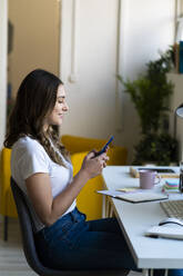 Smiling female entrepreneur using smart phone while sitting on chair at office - GIOF09819