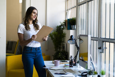 Smiling businesswoman holding clipboard while looking away in office - GIOF09832