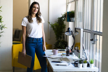 Young businesswoman holding clipboard while standing at office - GIOF09834