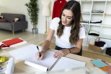 Smiling female fashion designer drawing sketch in book at studio - GIOF09841