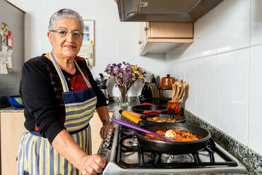 Ältere Hausfrau brät Fischsteaks in der Pfanne, während sie in der Küche ein typisches katalanisches Kabeljau-Gericht zubereitet und dabei in die Kamera schaut - ADSF18064