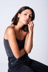 Full length young slim brunette in casual outfit sitting on stool against white background and looking at camera - ADSF18067