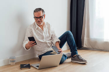 Full length senior male in casual outfit and eyeglasses sitting on floor with laptop and notebook while browsing modern mobile phone - ADSF18068