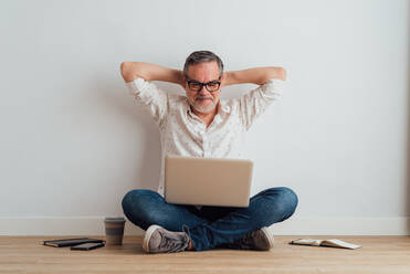 Full body delighted pleased mature male wearing casual clothes sitting on floor with contemporary laptop on laps - ADSF18069