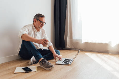 Nachdenklicher älterer Mann, der mit gekreuzten Beinen auf dem Parkett sitzt, ein Netbook auf dem Schoß und eine Tasse Kaffee zum Mitnehmen in der Hand, während er in Gedanken versunken ist - ADSF18070