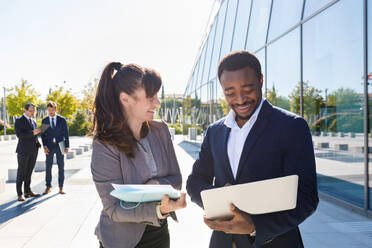 Focused young female manager in suit showing documents to African American male colleague standing near contemporary building with laptop on sunny day - ADSF18075