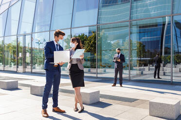 Unrecognizable young man and woman in formal suits and medical masks examining documents while working together on street using laptop near modern business building - ADSF18077