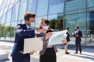 Unrecognizable young man and woman in formal suits and medical masks examining documents while working together on street using laptop near modern business building - ADSF18078