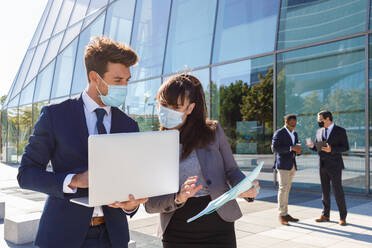 Unrecognizable young man and woman in formal suits and medical masks examining documents while working together on street using laptop near modern business building - ADSF18079