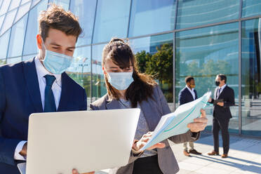 Unrecognizable young man and woman in formal suits and medical masks examining documents while working together on street using laptop near modern business building - ADSF18080