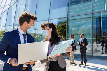 Unrecognizable young man and woman in formal suits and medical masks examining documents while working together on street using laptop near modern business building - ADSF18081