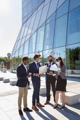 Full body of anonymous diverse coworkers working on laptop in formal suit and medical mask using laptop while standing on street - ADSF18082