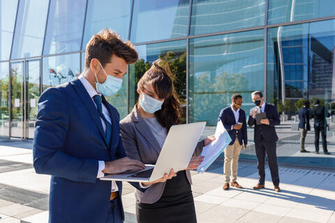 Unrecognizable young man and woman in formal suits and medical masks examining documents while working together on street using laptop near modern business building stock photo