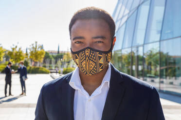 Happy young African American male entrepreneur with mask in classy suit standing on street neat contemporary business building and looking at camera - ADSF18087