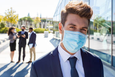 Serious young businessman with stylish hairstyle in formal suit and medical mask standing near glass facade of modern building and looking away during work with unrecognizable colleagues - ADSF18093