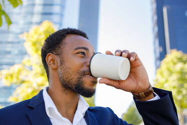 Niedriger Winkel des positiven jungen afroamerikanischen männlichen Managers in formellem Outfit, der während einer Pause an einem sonnigen Tag Kaffee zum Mitnehmen auf der Straße trinkt - ADSF18113