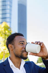 Niedriger Winkel des positiven jungen afroamerikanischen männlichen Managers in formellem Outfit, der während einer Pause an einem sonnigen Tag Kaffee zum Mitnehmen auf der Straße trinkt - ADSF18114