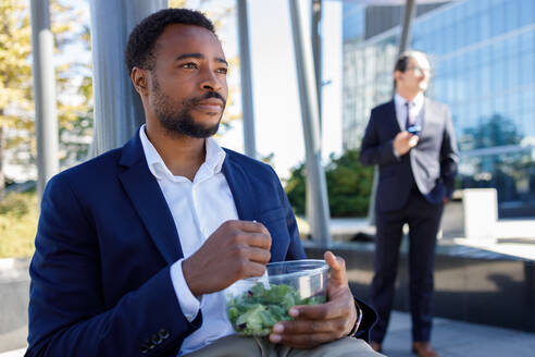 Nachdenklicher junger schwarzer Unternehmer im formellen Anzug isst gesunden Salat zum Mitnehmen, während er an einem sonnigen Tag eine Pause auf der Straße macht - ADSF18118