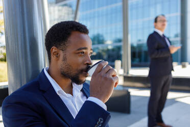 Seitenansicht eines konzentrierten jungen afroamerikanischen männlichen Unternehmers im eleganten Anzug, der auf einer Straße in der Nähe moderner Wolkenkratzer einen Kaffee zum Mitnehmen trinkt und wegschaut - ADSF18119