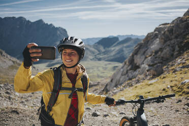 Lächelnde Radfahrerin, die ein Selfie mit ihrem Mountainbike am Berg macht, Nationalpark Picos de Europa, Kantabrien, Spanien - DMGF00371