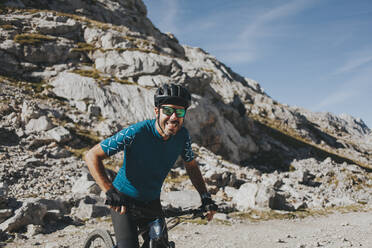 Fröhlicher männlicher Radfahrer auf dem Fahrrad gegen einen Berg an einem sonnigen Tag, Nationalpark Picos de Europa, Kantabrien, Spanien - DMGF00373