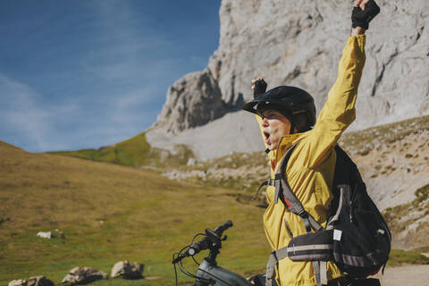 Excited female cyclist with arms raised shouting at Picos de Europa National Park on sunny day, Cantabria, Spain stock photo