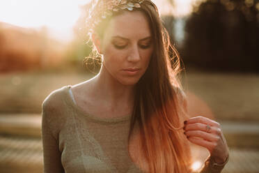 Crop serene female wearing transparent sweater standing in lush green park in evening sunlight and looking down pensively - ADSF18142