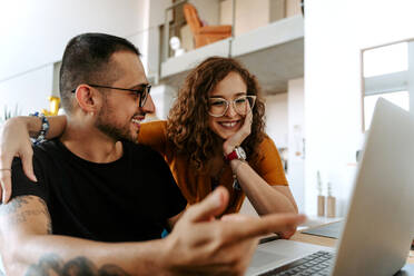 Cheerful young diverse couple in casual clothes and eyeglasses holding hands while sitting together on sofa and having video call on laptop - ADSF18158