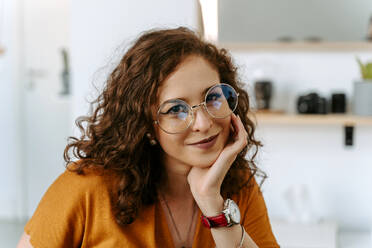 Positive junge Frau mit lockigem rotem Haar in Freizeitkleidung und trendiger Brille, die lächelnd am Tisch sitzt, die Hand am Kinn hat und in die Kamera schaut - ADSF18161