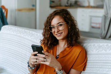 Concentrated young female with curly hair in casual clothing surfing internet while resting on sofa in bright room - ADSF18169