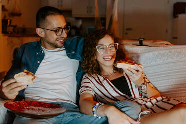 Front view of young multiracial couple in casual clothes cuddling and eating pizza while sitting together on couch and watching movie on laptop - ADSF18172