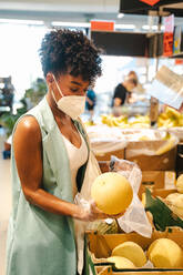 Seitenansicht einer afroamerikanischen Kundin mit Schutzmaske und Handschuhen, die während des Einkaufs im Supermarkt während der Coronavirus-Pandemie reife Wassermelonen aus einer Kiste pflückt - ADSF18173