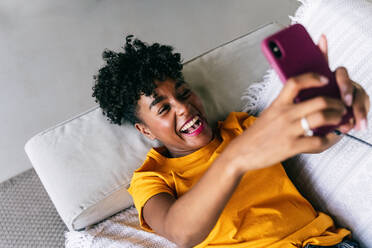 Side view of excited African American millennial female in casual outfit taking selfie with mobile phone and laughing while resting on sofa in modern apartment - ADSF18176