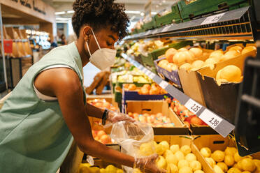 Side view of young African American female in protective mask and gloves choosing red apples while visiting grocery store during coronavirus pandemic - ADSF18178