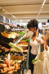 Side view of young African American female in protective mask and gloves choosing red apples while visiting grocery store during coronavirus pandemic - ADSF18179