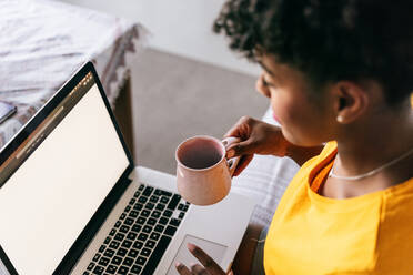 Fokussierte junge afroamerikanische Freiberuflerin mit einer Tasse Kaffee in der Hand, die auf dem Sofa sitzt und auf ihrem Laptop surft, während sie zu Hause an einem Projekt arbeitet - ADSF18181