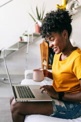 Focused young African American female freelancer with cup of coffee in hand sitting on sofa and browsing laptop while working remotely on project at home - ADSF18182