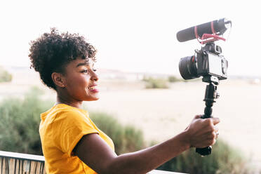 Cheerful young African American female blogger holding selfie stick with camera and recording video for vlog while standing on terrace against blurred nature during summer travel - ADSF18185