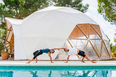 Schlanke Partner in Sportkleidung, die in Camatkarasana-Pose stehen, während sie in der Nähe des Swimmingpools Yoga praktizieren - ADSF18201