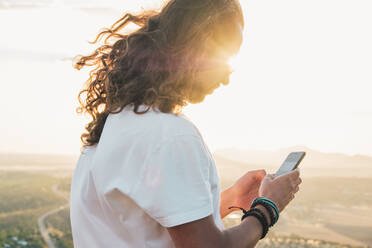 Side view crop calm male with long curly hair browsing contemporary mobile phone while enjoying sunny evening on hilltop against blurred scenic grassland - ADSF18214