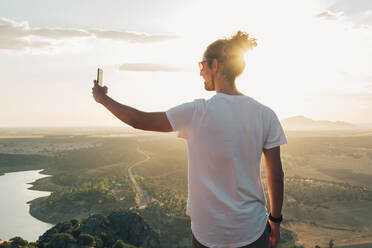 Rückenansicht anonymer Reisender in Freizeitkleidung, der auf einer Bergkuppe steht und mit seinem Smartphone Fotos von einem weitläufigen, grasbewachsenen Tal im Sonnenlicht macht - ADSF18216