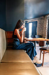 Side view of serene young brunette female in elegant outfit sitting on table drinking coffee in room with stylish interior - ADSF18220