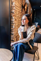 Seitenansicht einer fröhlichen jungen brünetten Frau in elegantem Outfit, die auf einem Stuhl vor einem Fenster sitzt und Kaffee in einem Raum mit stilvollem Interieur trinkt - ADSF18223