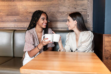 Fröhliche junge gemischtrassige Frauen in trendigen Outfits, die Kaffee trinken und fröhlich plaudern, während sie in einer modernen Cafeteria Zeit miteinander verbringen - ADSF18225