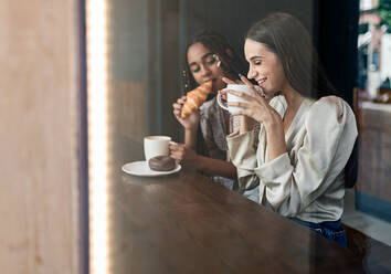 Durch das Fenster Blick auf fröhliche Millennial diverse Freundinnen plaudern glücklich, während an der Theke im Café sitzen und genießen heißen Kaffee mit Gebäck - ADSF18226